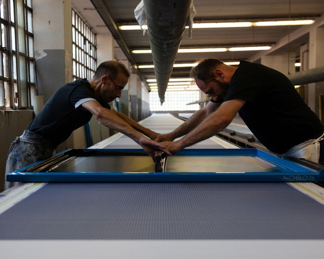 The process begins with a roll of white silk stretched out on a ten-metre bench. Two workers move in unison along its length, carrying the printing frame.